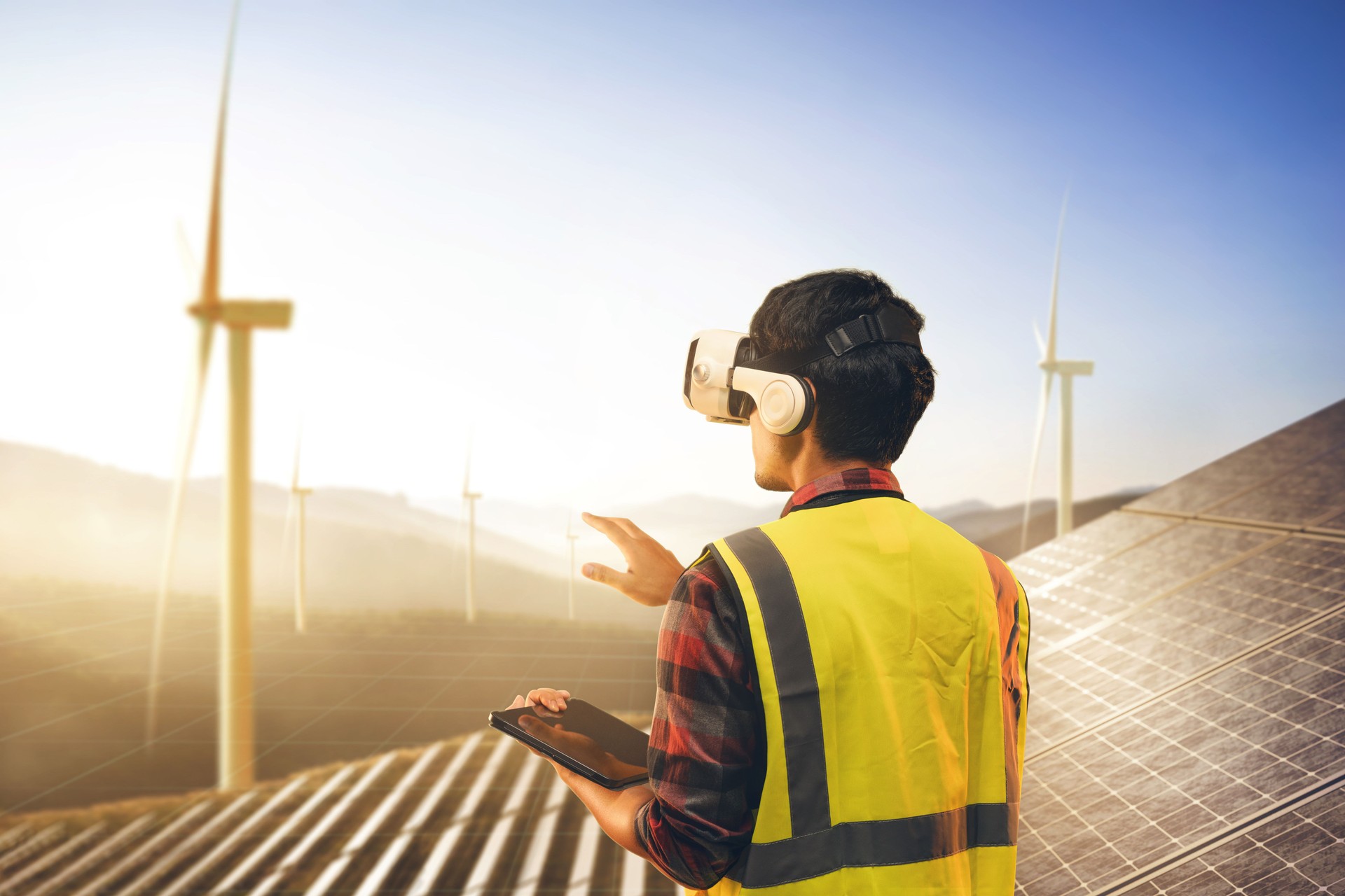 Engineer working with technology metaverse with VR headset for plan or checking operation of field of photovoltaic solar panels and wind turbine power plant on the mountain.