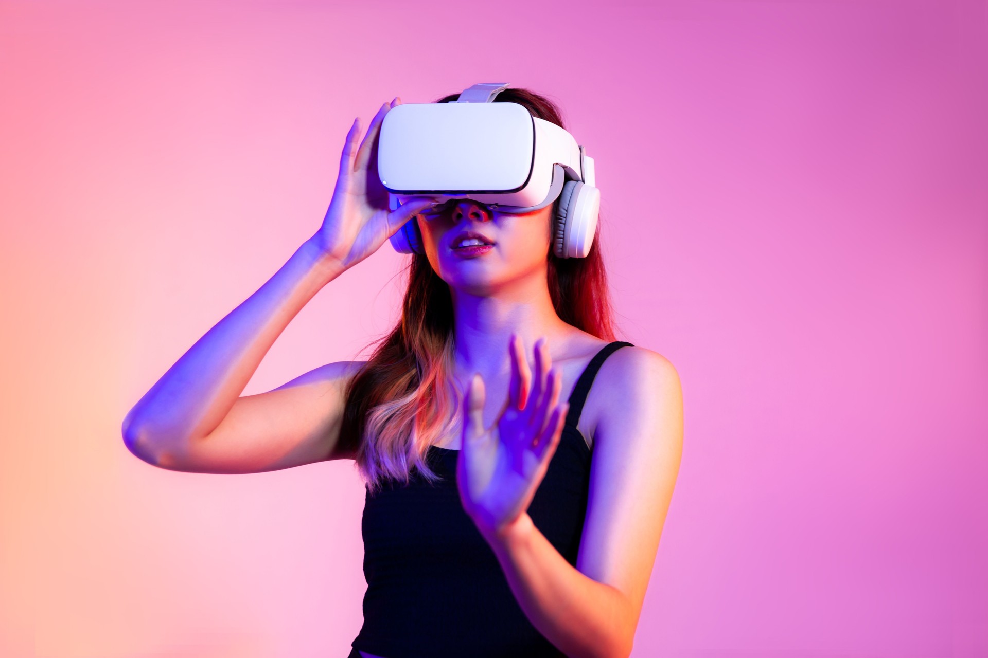 Metaverse concept young asian woman in black tank top wearing white vr headset hand touching watching and playing on the pink screen background."t